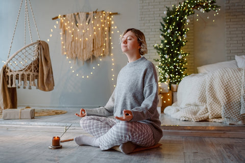Beautiful young woman sitting on the floor relaxing in lotus position with eyes closed meditating in Christmas decorated home. Mental healthcare, care of yourself during holiday